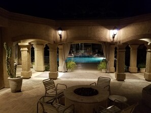 Un patio al aire libre con columnas de piedra, una fogata y una piscina al fondo.