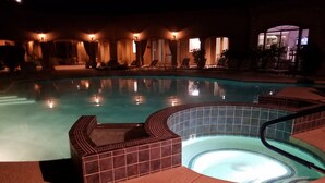 Un área de piscina nocturna con un jacuzzi, sillas de descanso y un deck de azulejos.