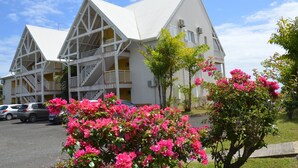 A white building with a triangular roof and balconies. There are cars parked in front and pink flowers in the foreground. - Caribbean Village, holiday apartment, Guadeloupe, 97180 (Sainte-Anne)