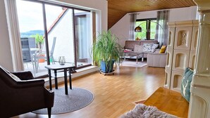A spacious living room with a large window, a round coffee table, a sofa, and a potted plant.