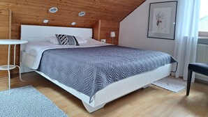 A modern bedroom with a wooden ceiling, a white bed with a grey quilt, a small white bedside table, and a framed picture on the wall.