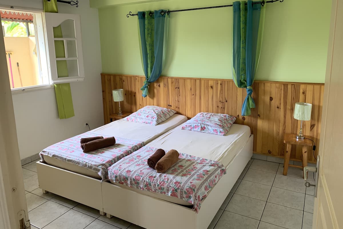 Cama doble con ropa de cama floral y dos almohadas, cabecera de madera, mesita de noche con lámpara y ventana con cortinas.