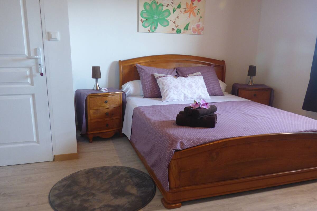 Un dormitorio con una cama de madera, dos mesitas de noche, un tapete redondo y un adorno floral en la pared.
