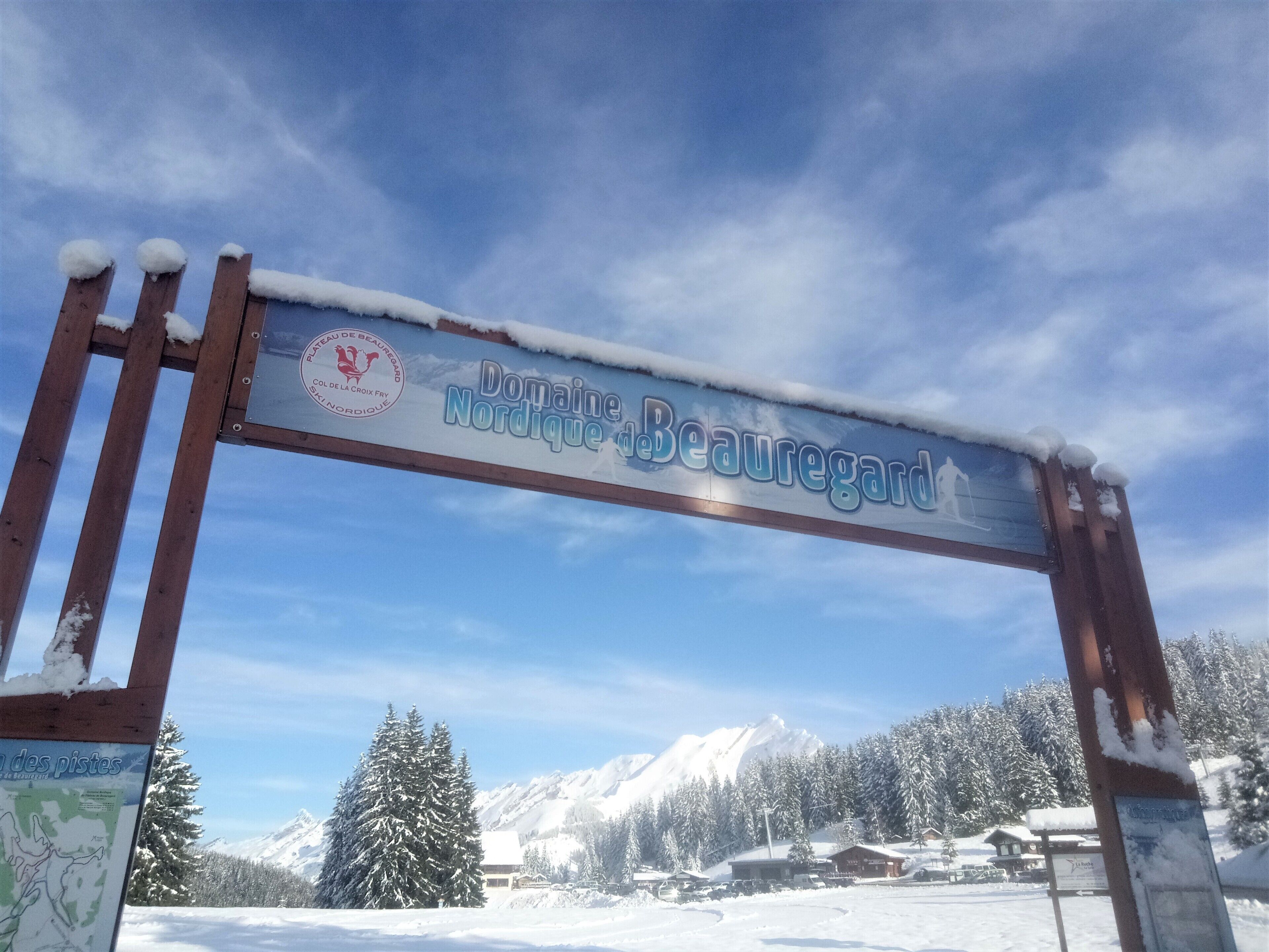 A wooden sign with the text "Domaine Nordique Beauregard" and a logo, set against a snowy landscape with mountains in the background.