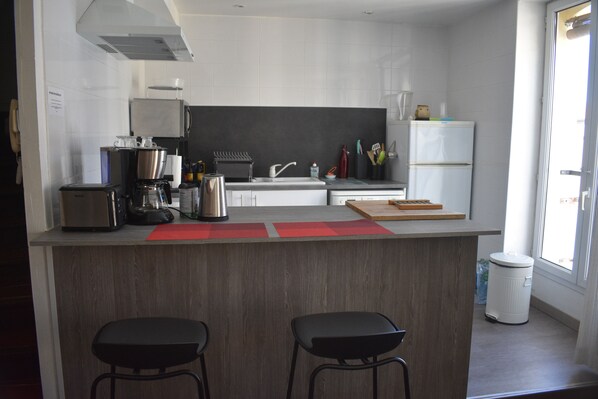 Una cocina moderna con una isla central, dos taburetes de bar, un refrigerador de acero inoxidable y un ventanal que proporciona luz natural.