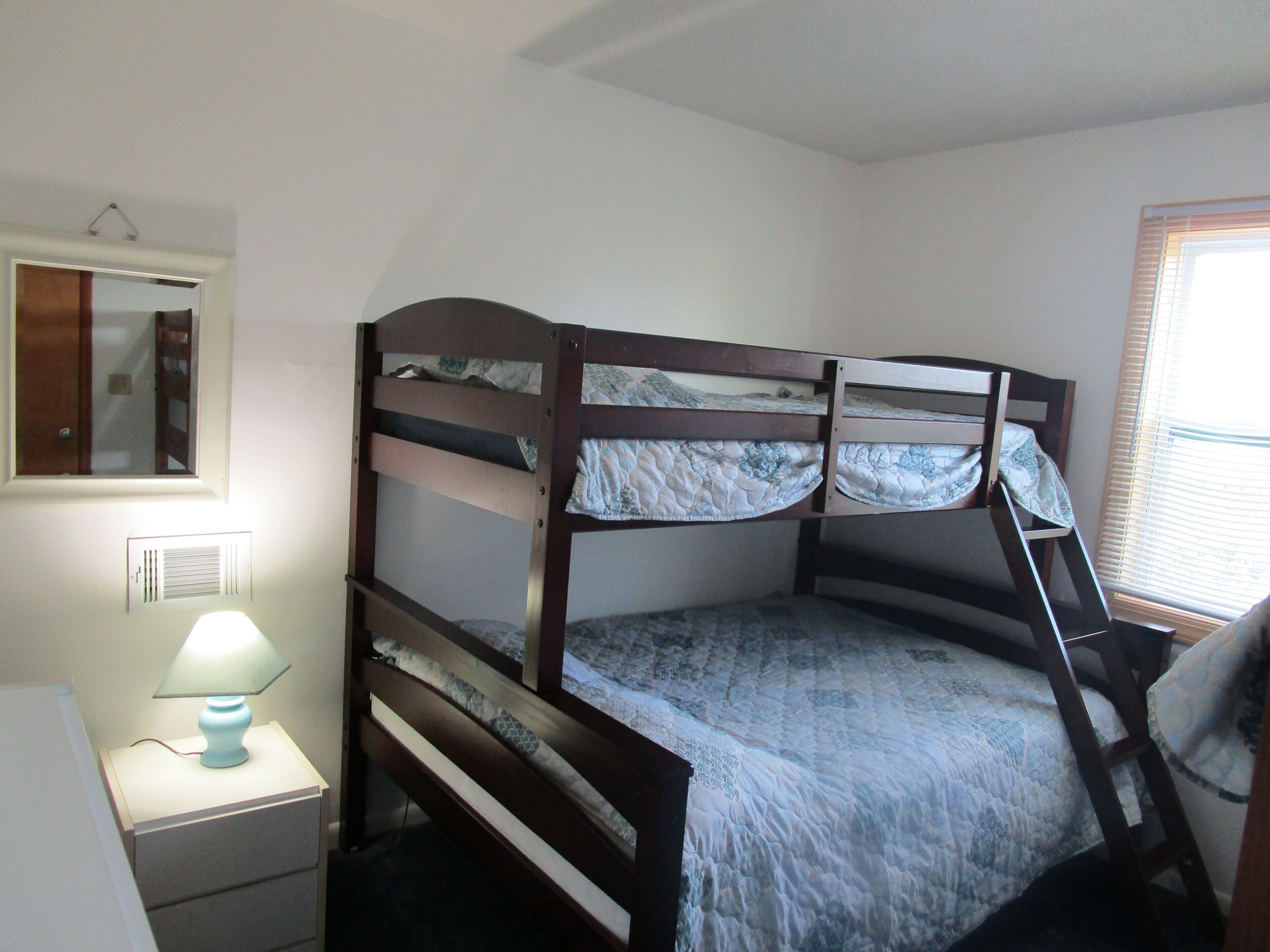 A bunk bed with a ladder, a bedside table with a lamp, a mirror, and a window with blinds.