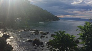 A coastal landscape with a rocky shoreline, calm sea, and a forested hill. - Camping do Bené (Angra dos Reis)