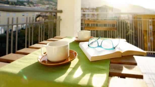 A balcony with a table set for two, featuring a cup, saucer, book, and glasses.