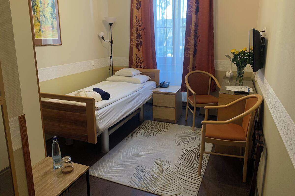 A small, tidy room with a single bed, a desk with a chair, a lamp, a TV, and a window with curtains.