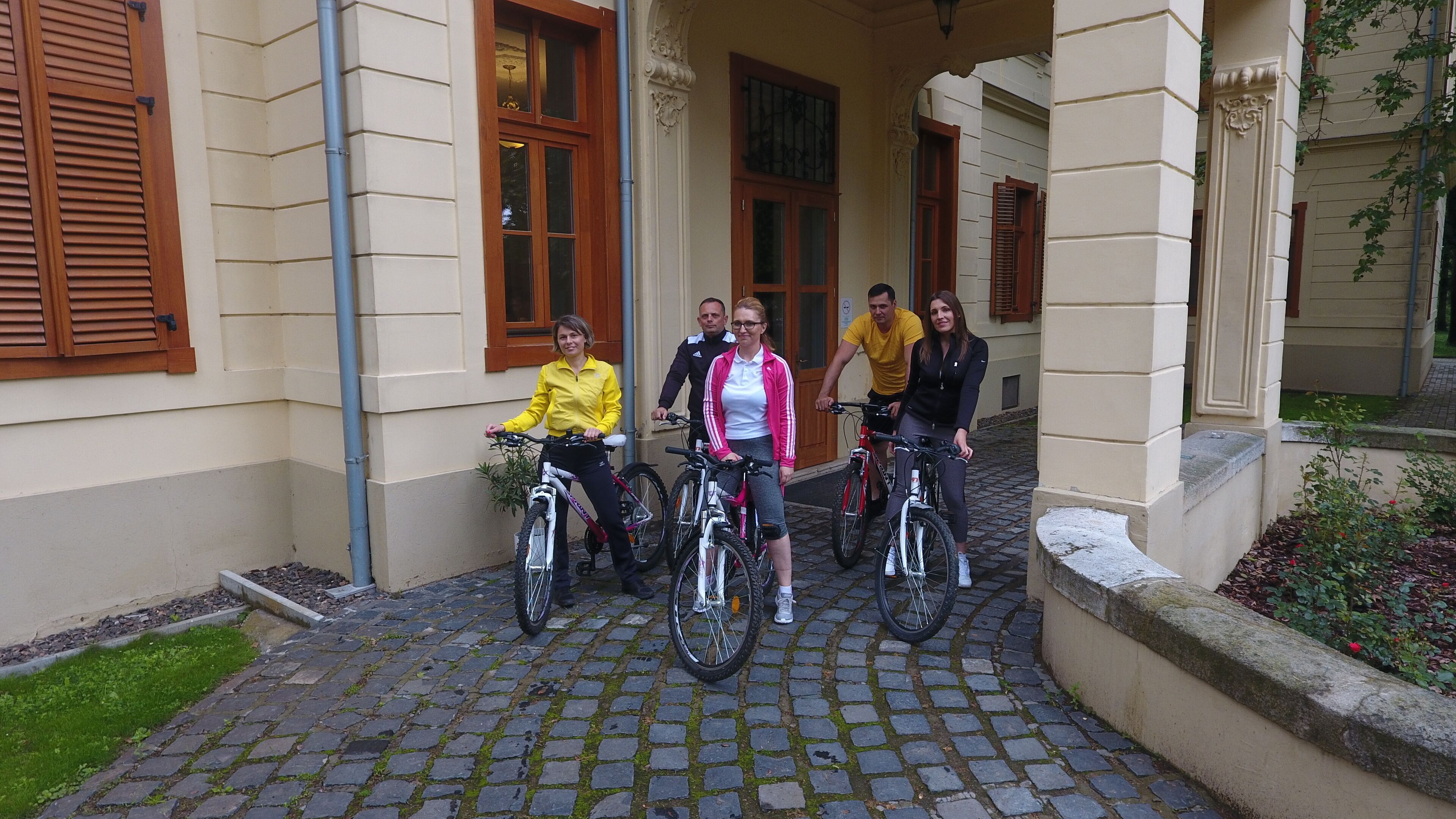 A group of people on bicycles in front of a building with a cobblestone entrance and wooden shutters.