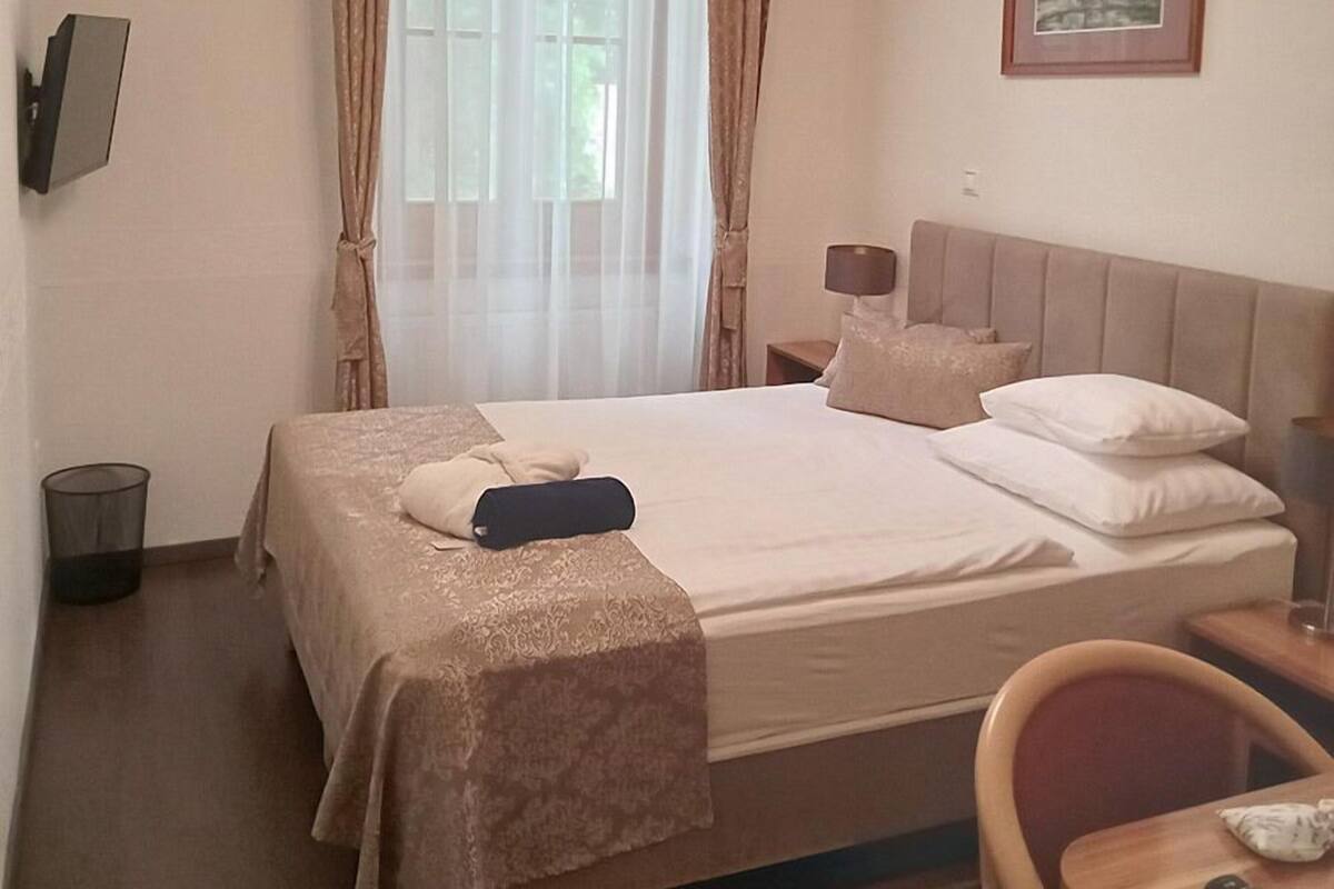 A neatly made bed with a beige bedspread and pillows, a bedside lamp, a small table with a remote control, and a chair.
