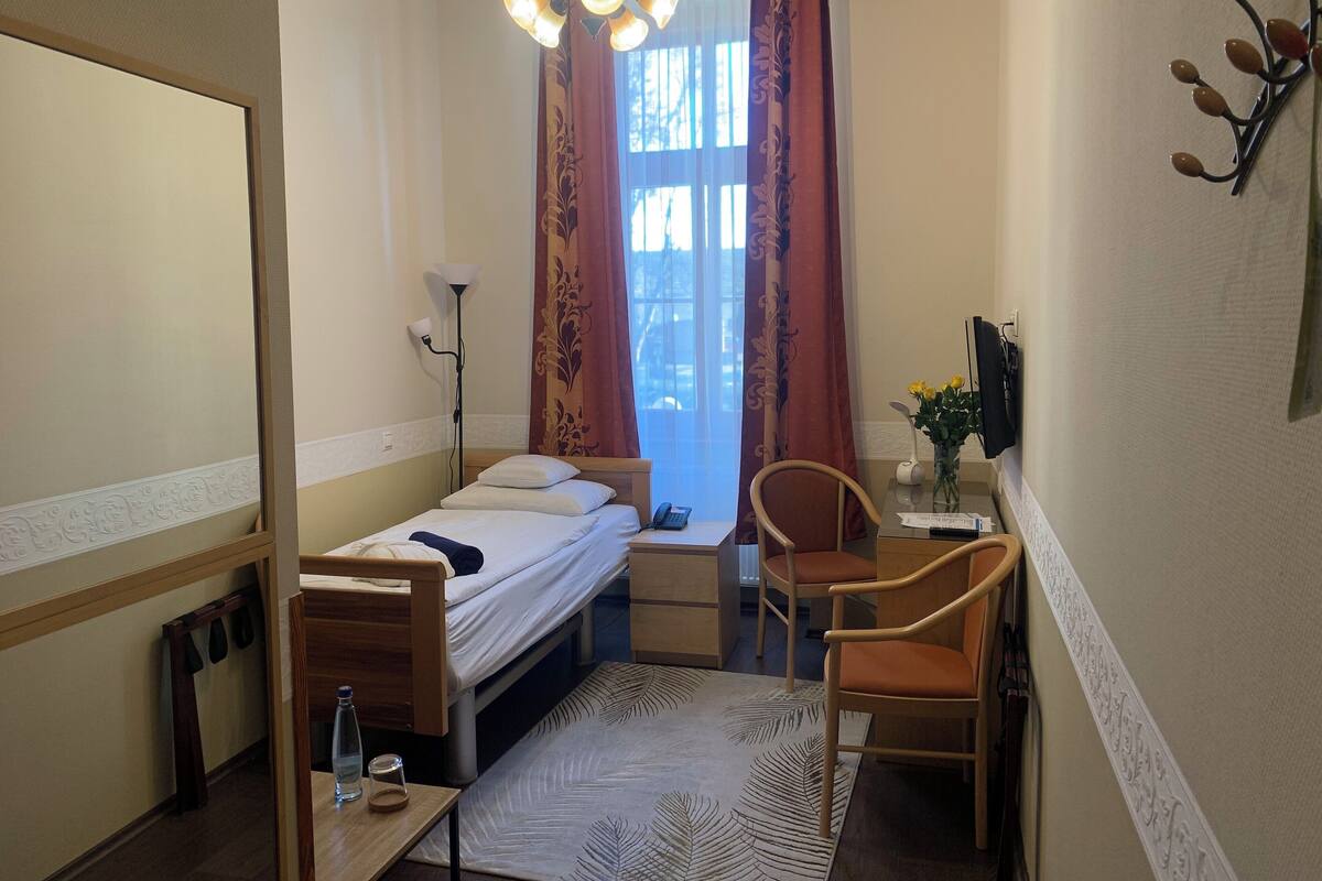 A small, neatly arranged bedroom with a single bed, a desk, a chair, a bedside table with a lamp, and a window with curtains.