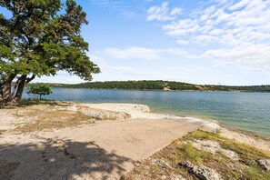 Une zone au bord du lac, avec une plage de sable, un grand arbre et, au loin, une colline parsemée de bâtiments.
