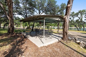 Un espace pique-nique abrité, équipé de tables et de chaises, entouré d’arbres et bordé par un plan d’eau.