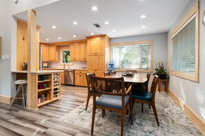 A modern kitchen with wooden cabinets, a dining area with a table and chairs, and a large window with blinds.