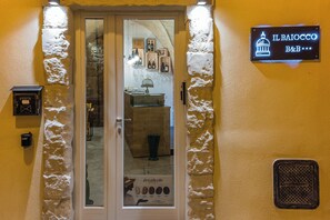 A hotel entrance with a glass door, a sign above indicating the name 'IL BAIOCCO B&B', and a display case with wine bottles inside. - Il Baiocco -Ibla Charme Rooms (Ragusa)