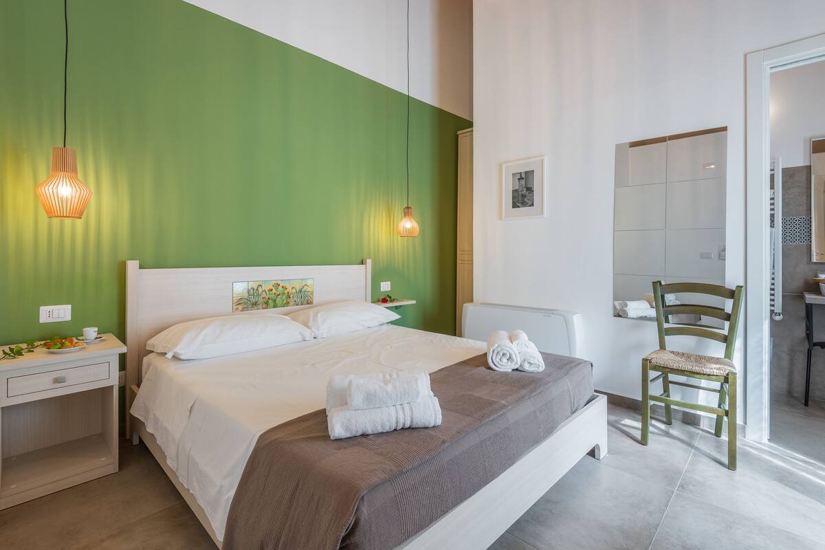 A neatly made bed with white linens, a brown throw blanket, and neatly folded towels on the side. There is a wooden chair with a cushion and a small table with a cup and a plate of fruit.