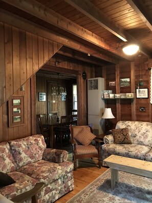 A cozy cabin interior with wooden walls, a dining area with chairs and a table, and two floral-patterned sofas.