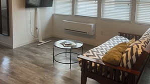 Un salon moderne avec un plancher en bois, un canapé agrémenté de coussins à motifs, une table basse en verre, un ventilateur de plafond et un téléviseur fixé au mur.
