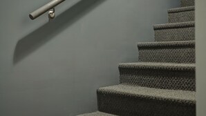 Un escalier dont les marches sont recouvertes de moquette, avec une rampe métallique, le tout adossé à un mur sobre.
