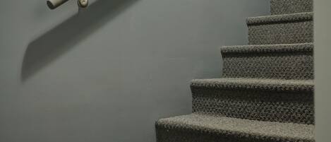A staircase with carpeted steps and a metal handrail against a plain wall.
