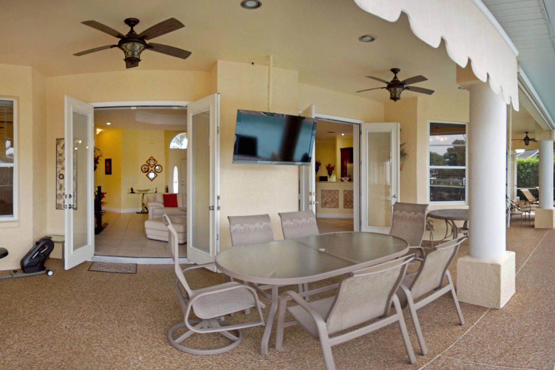 A covered outdoor dining area with a round table and chairs, ceiling fans, and a TV mounted on the wall.