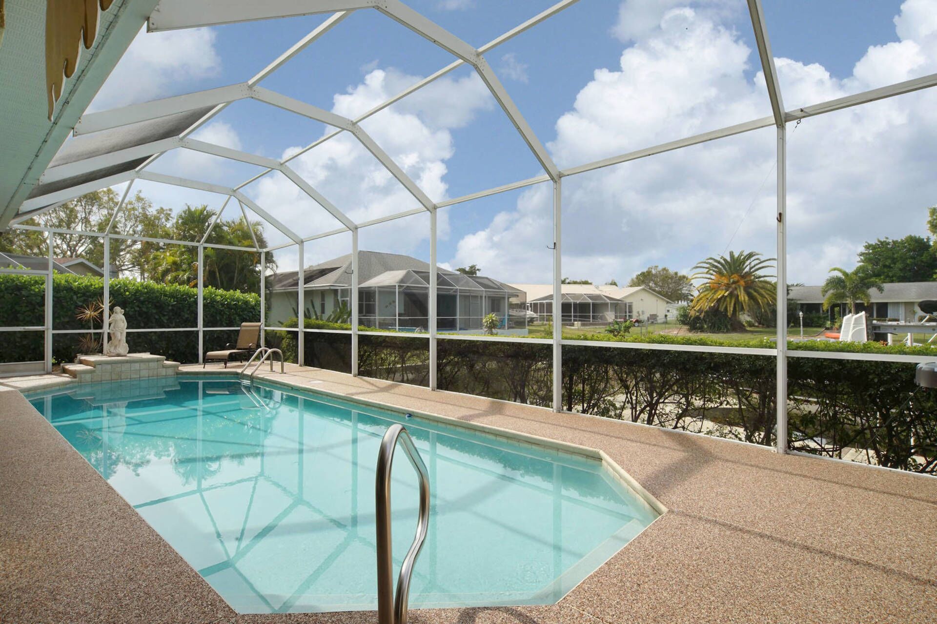 A covered swimming pool with a glass barrier, surrounded by a white metal frame, and a view of houses and greenery in the background.