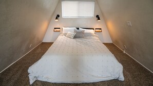 A small, triangular attic bedroom with a single bed, two wall-mounted reading lamps, and a window with blinds. - Stylish A-Frame (Eden)