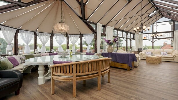 Ein Wintergarten mit einem Glastisch, Holzstühlen, einer Couch und einer Hängelampe.
