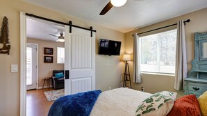 Un dormitorio con cama, televisor, ventilador de techo, ventana con cortinas y puerta que da a otra habitación.
