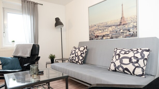 A modern living room with a grey sofa, patterned cushions, a glass coffee table, a black armchair, a floor lamp, and a large cityscape poster on the wall. - P1 - Large apartment at the gates of Kassel (Fuldatal)