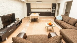 A modern living room with a flat-screen TV, a sofa, a dining table, and a kitchen in the background.