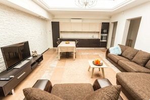 A modern living room with a flat-screen TV, a sofa, a dining table, and a kitchen in the background.