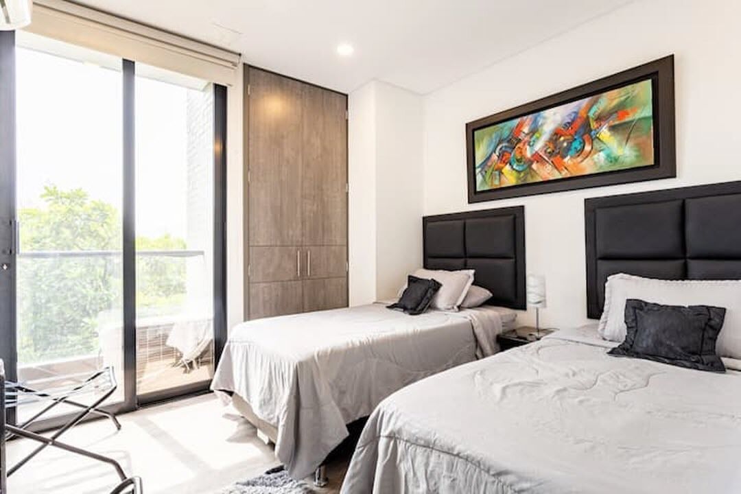 Habitación de hotel con dos camas, puerta corredera de vidrio a un balcón y un cuadro abstracto de colores en la pared.