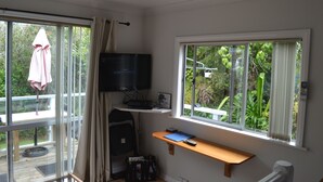 A room with a TV, a wooden shelf with a notebook, a window with curtains, and a view of greenery outside. - The Lighthouse (Torbay)