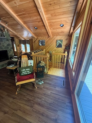 A cabin interior with a wooden ceiling, stone fireplace, wooden furniture, and a view through large windows.