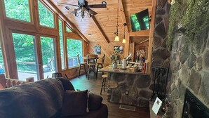A wooden interior with a stone fireplace, a wooden ceiling, and large windows. - Beautiful 2 BR cabin with a wall of windows overlooking the deck and stream. (Dayton)