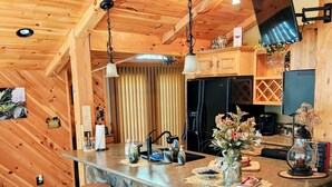 A kitchen with a stone island, wooden bar stools, and a TV mounted on the wall. - Beautiful 2 BR cabin with a wall of windows overlooking the deck and stream. (Dayton)