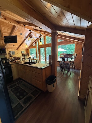 A wooden cabin interior with a kitchen, dining area, and large windows.