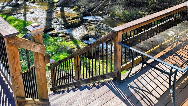 A wooden deck with a railing overlooking a forested area with a stream. - Beautiful 2 BR cabin with a wall of windows overlooking the deck and stream. (Dayton)
