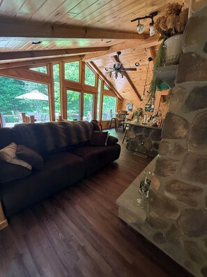 LâintĂ©rieur chaleureux dâun chalet avec une cheminĂ©e en pierre, un plafond en bois et de grandes fenĂȘtres donnant sur un jardin.
