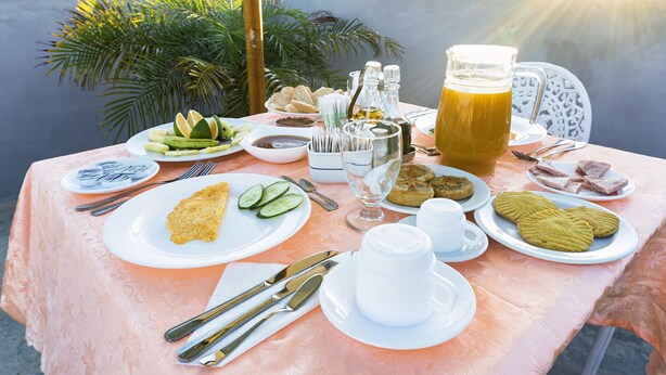 Ein Tisch ist reichhaltig mit Frühstück gereicht, darunter Eier, Gurkenscheiben und Gebäck, sowie ein Saftkrug und Beilagen.