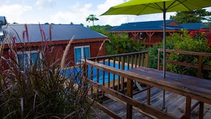 Une terrasse en bois avec une piscine, entourée d’une végétation luxuriante et d’un bâtiment rouge coiffé d’un toit bleu.
