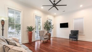 A spacious living room with hardwood flooring, a ceiling fan, a large window, and a flat-screen TV mounted on the wall. - Historic Mini-Mansion on Canal St. Walk to (New Orleans)