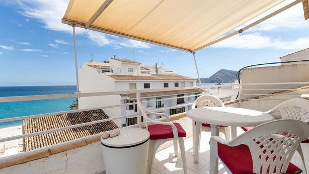 Varanda com cadeiras e mesa de plástico branco, com vista para a praia e edifícios.