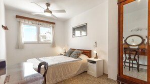 Quarto com cama, mesas de cabeceira, escrivaninha, cadeira, ventilador de teto, janela com cortinas, quadro emoldurado e espelho.