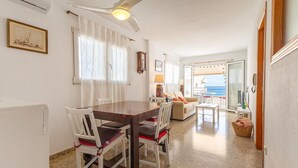 Uma área de refeições com mesa e cadeiras, um sofá, um abajur e vista para o mar através de uma porta de vidro.