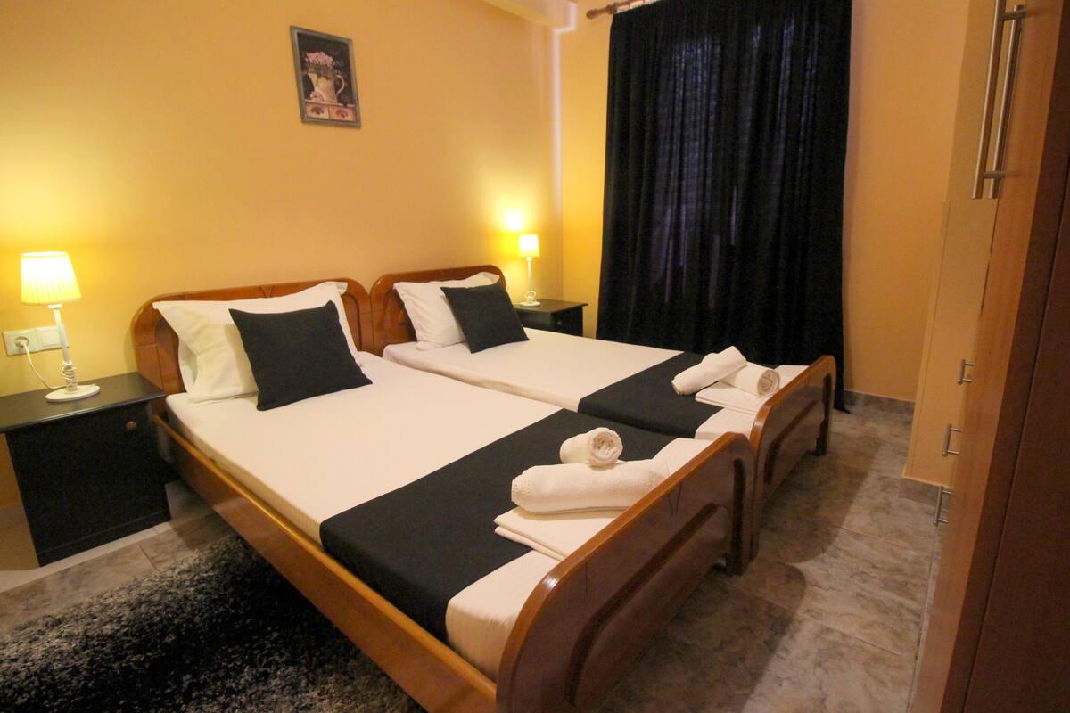 A hotel room with two beds, a dark curtain, wooden headboards, and a small nightstand with a lamp.