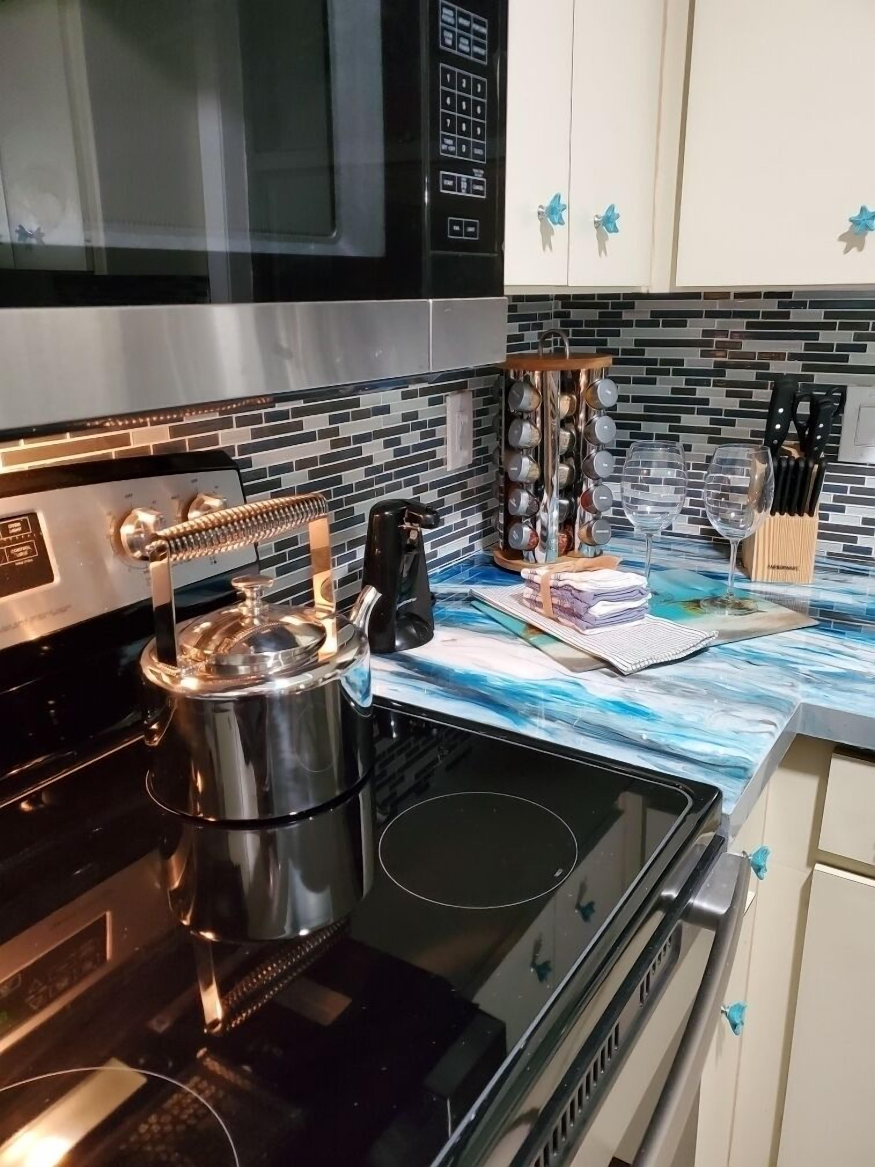 Une cuisine équipée d’une cuisinière en acier inoxydable, d’une bouilloire et d’un four à micro-ondes. On y trouve des placards avec des poignées bleues et un pare-feu en carreaux.