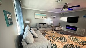 Un salon comprenant un canapé, une table basse et un tapis à motifs. Un téléviseur est fixé au mur et un ventilateur de plafond est installé.